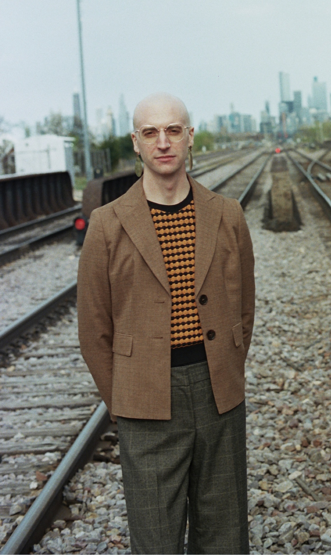 A bald white person with glasses and earrings wearing neutral colors in a blazer and dress paints stands among train tracks leading to the Chicago skyline 